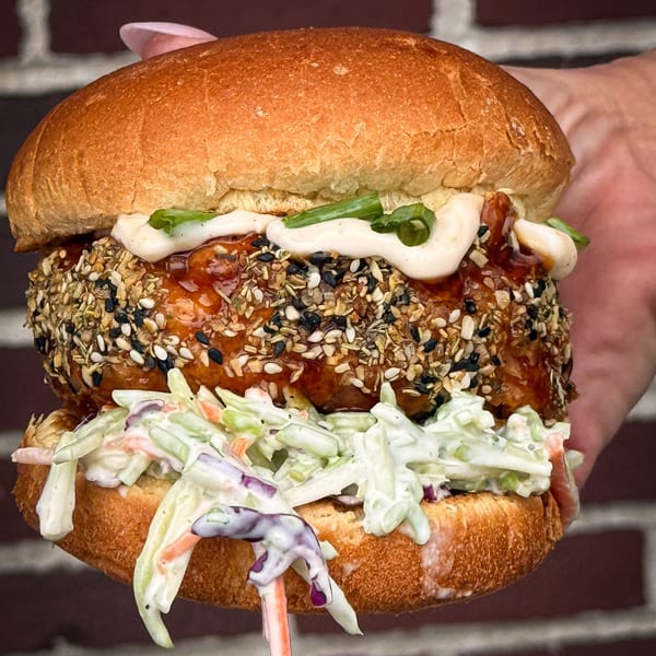 Hand holding a sesame-crusted chicken sandwich with coleslaw, sauce, and green onions against a brick wall.