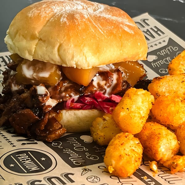 A vegan pulled pork burger with pickled cabbage and tater tots on branded paper.