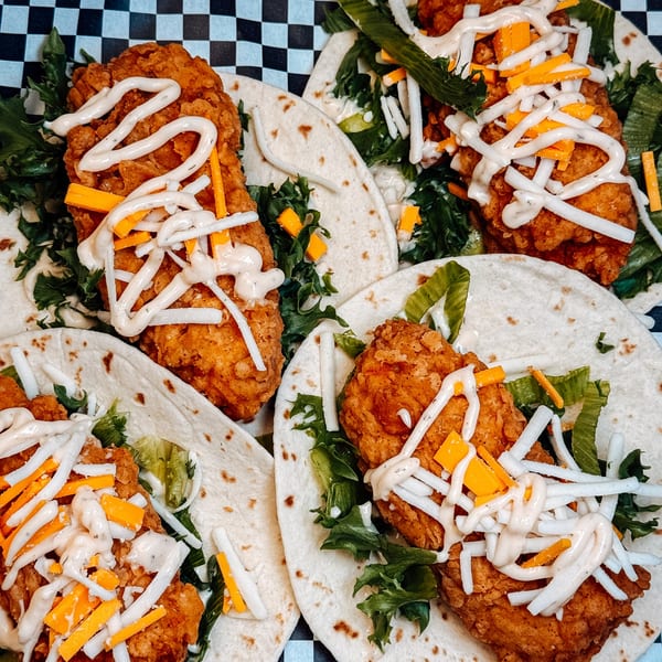 Crispy chicken tacos with lettuce, shredded cheese, and a creamy sauce on a checkered background.