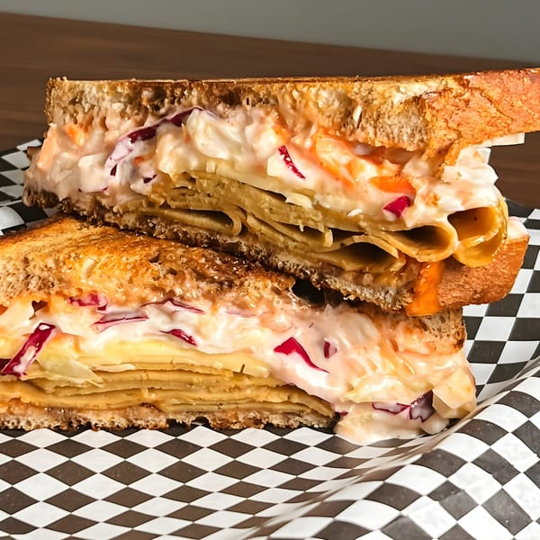 A vegan sandwich with layers of plant-based meat, cheese, and coleslaw on toasted bread.